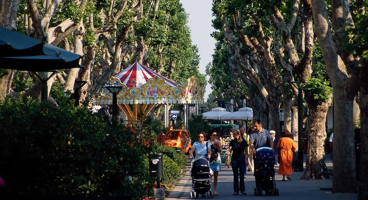 Passeggiata nel centro di Bellaria, con famiglie che spingono passeggini sotto alberi maestosi e un colorato carosello.