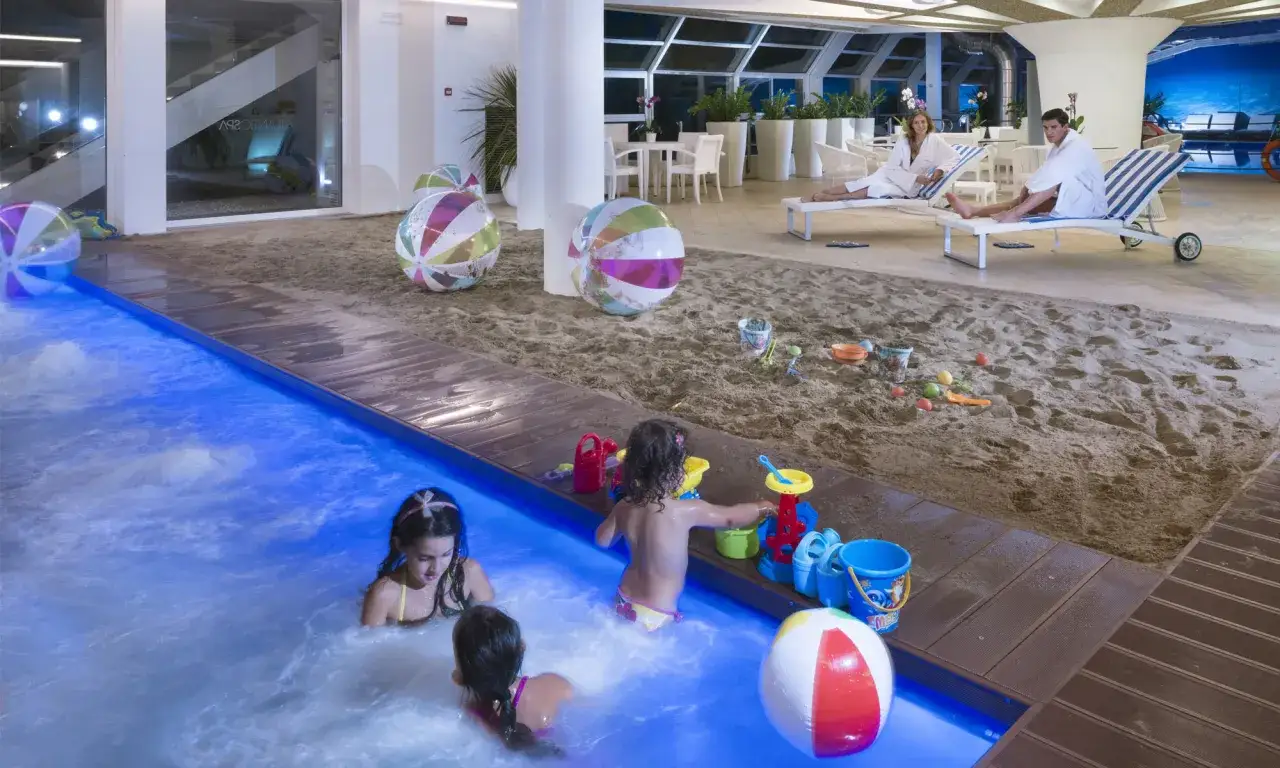 Bambini giocano in piscina e sulla sabbia in un ambiente che ricorda il bagno 110 Riccione, con palloni colorati e lettini.