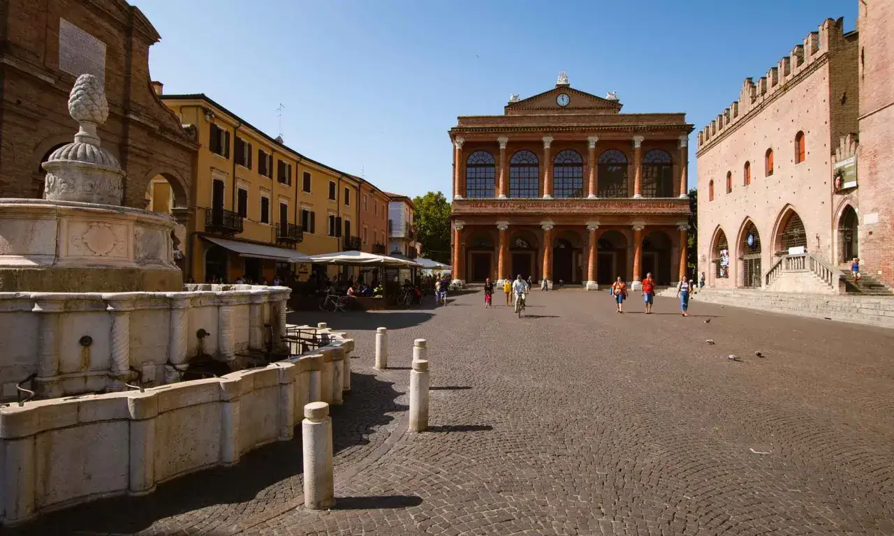 La piazza rimini, con la sua fontana storica e gli edifici antichi, &egrave; animata da persone che passeggiano e ciclisti sotto un cielo azzurro.