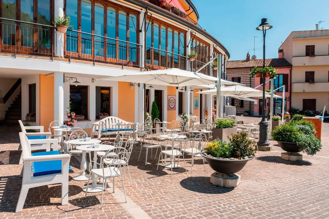 Tavolini all'aperto e sedie bianche davanti a una gelateria a Gabicce Mare, con ombrelloni aperti e piante verdi.