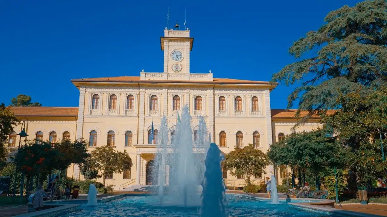 Fontana zampillante nella piazza Roosevelt di Cattolica, con edificio giallo e torre dell'orologio.