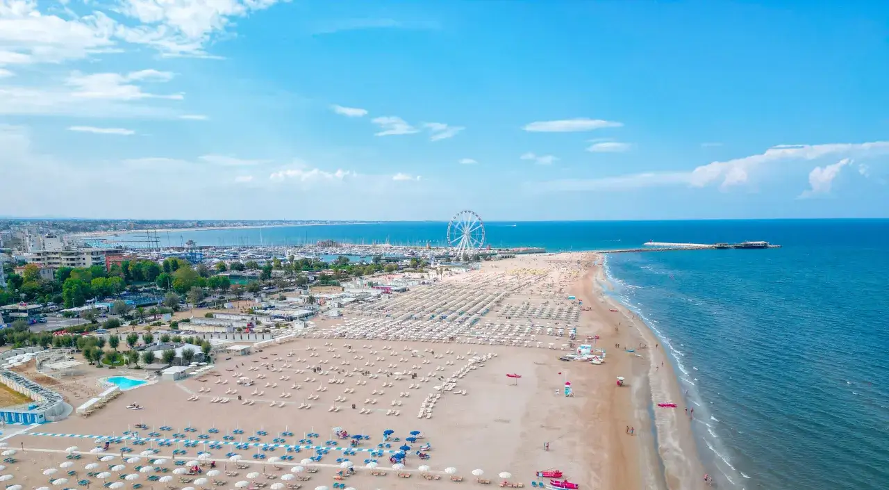 Spiaggia affollata a Rimini estate, con ombrelloni, lettini, mare azzurro e la ruota panoramica sullo sfondo.
