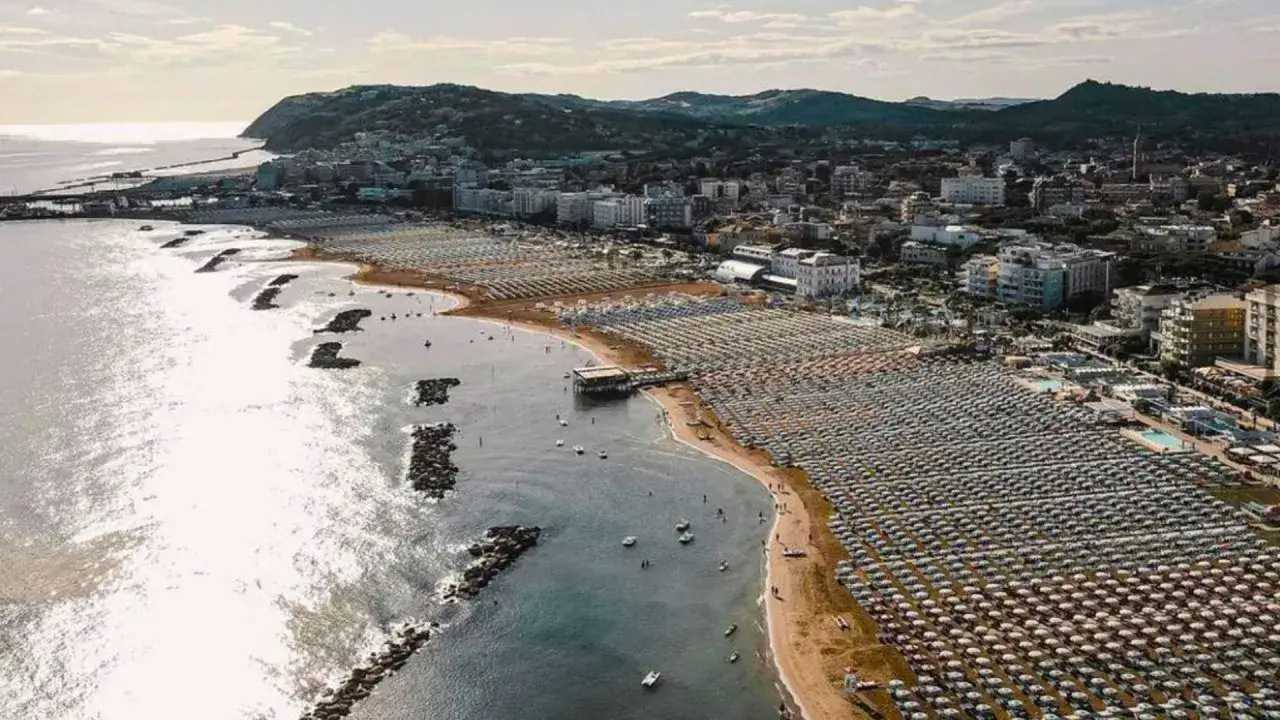 Vista aerea di una spiaggia affollata di ombrelloni a Cattolica, Emilia Romagna, con il mare scintillante e le colline sullo sfondo.