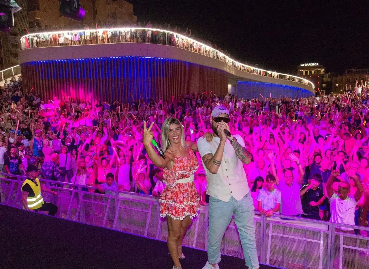 Una folla festante sotto luci rosa per la Notte Rosa a Rimini. Due artisti sul palco, un uomo canta e una donna sorride con un microfono.