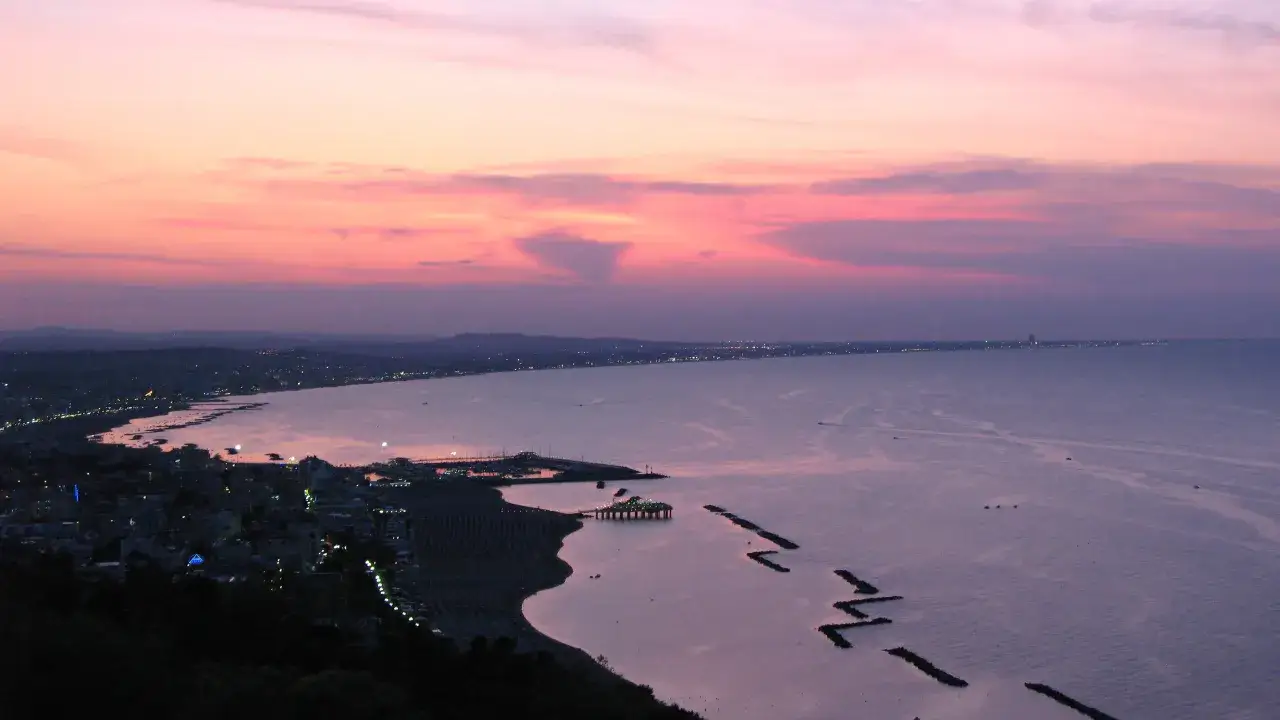 Panoramica Gabicce al tramonto, con cielo rosa e viola, mare calmo e costa illuminata.