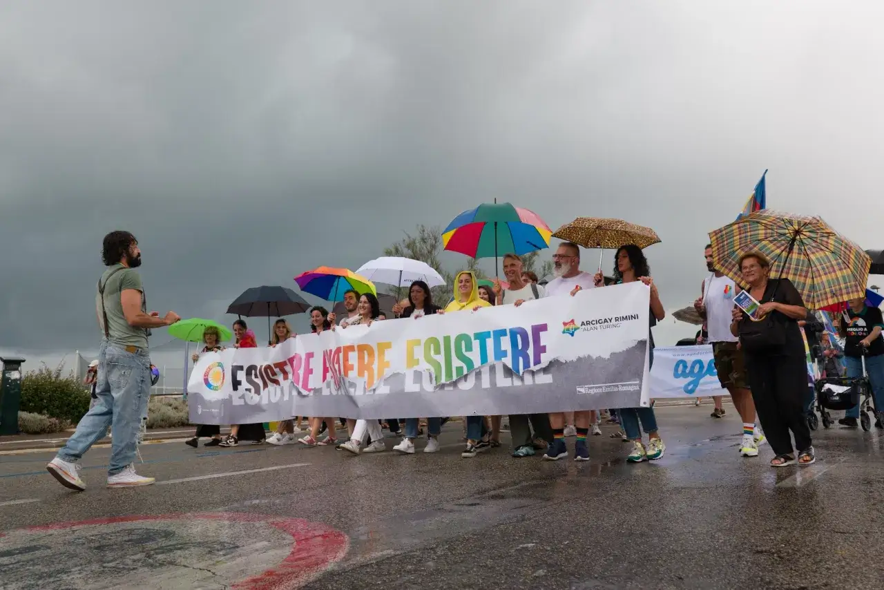 Persone con ombrelli colorati sfilano a Rimini Pride, tenendo uno striscione arcobaleno con la scritta 