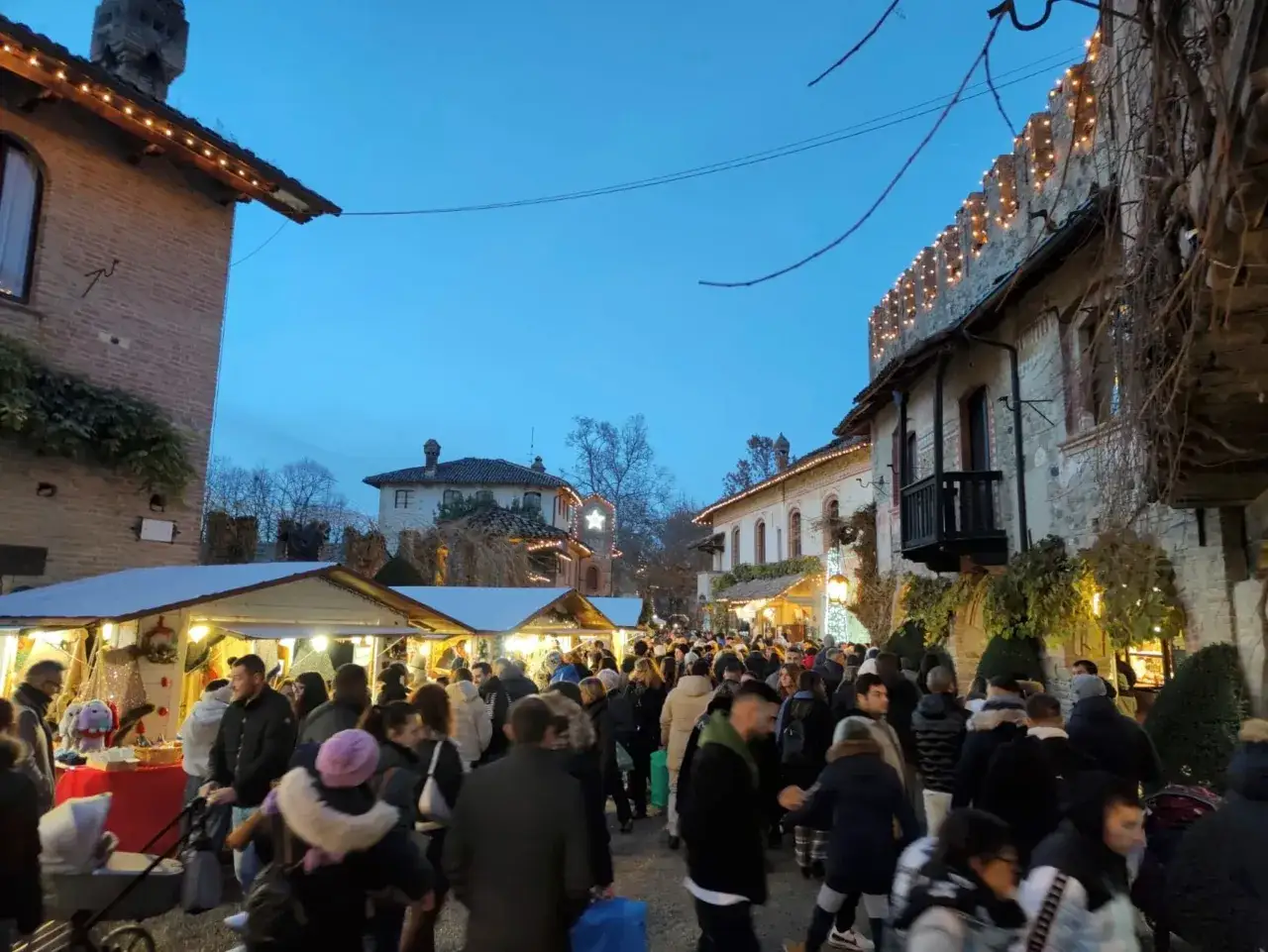 Folla ai mercatini di Natale in Romagna, tra bancarelle illuminate e antiche mura decorate con luci festive.