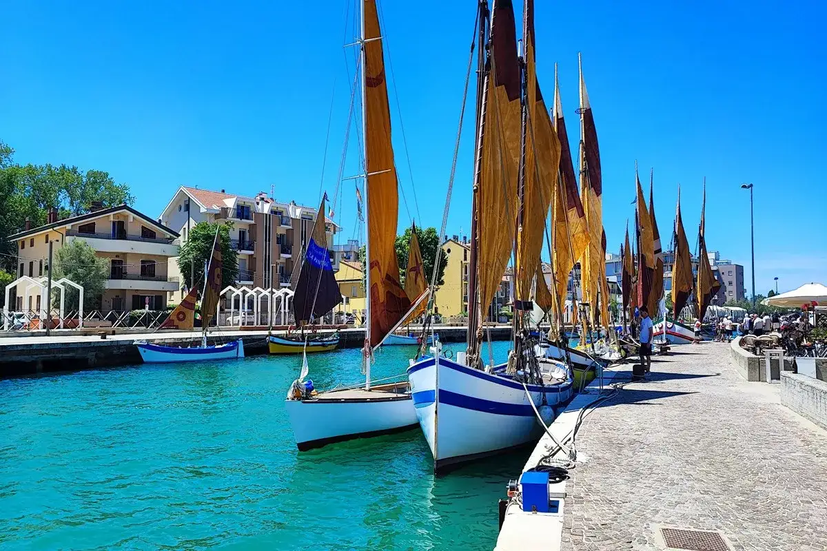 Vele colorate e barche tradizionali ormeggiate nel porto di Bellaria Igea, con edifici sullo sfondo e un cielo azzurro.