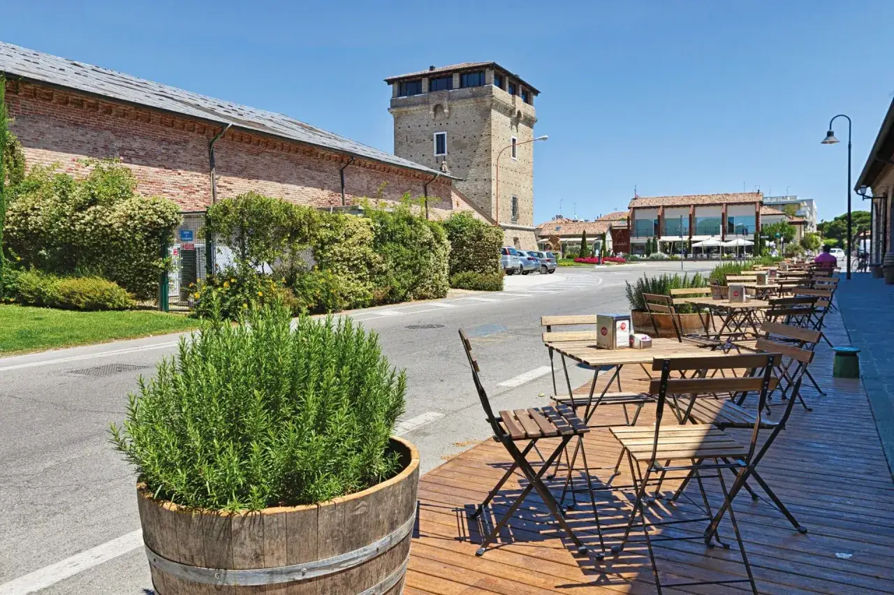 Davanti ai magazzini del sale di Cervia, tavolini e sedie in legno invitano a una pausa all'aperto, con un grande vaso di rosmarino a fare da cornice.