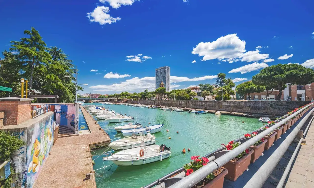 Il porto di Rimini, con barche ormeggiate in un canale turchese sotto un cielo azzurro e nuvoloso, alberi verdi e un grattacielo sullo sfondo.
