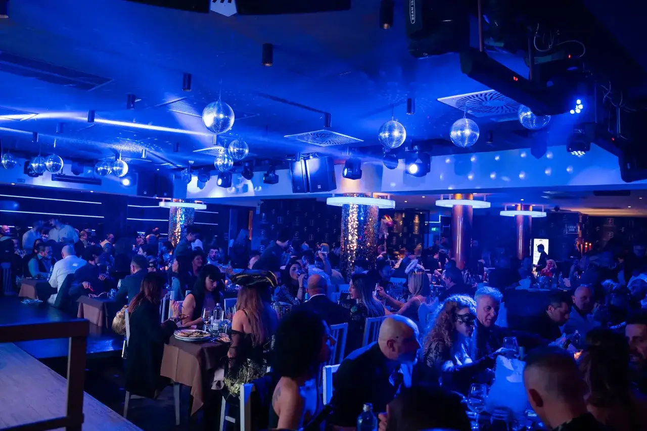 Atmosfera vibrante in un ristorante frontemare a Rimini, con luci blu, sfere da discoteca e gente che cena e festeggia.