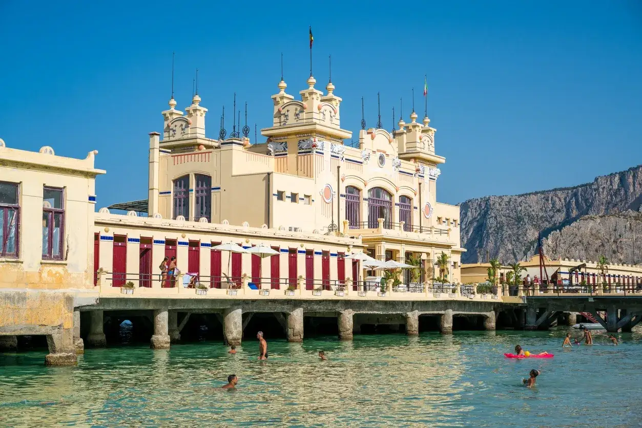 Edificio storico giallo con cabine rosse, mare cristallino e gente che nuota. Il primo stabilimento balneare in Italia, un'icona di Mondello.