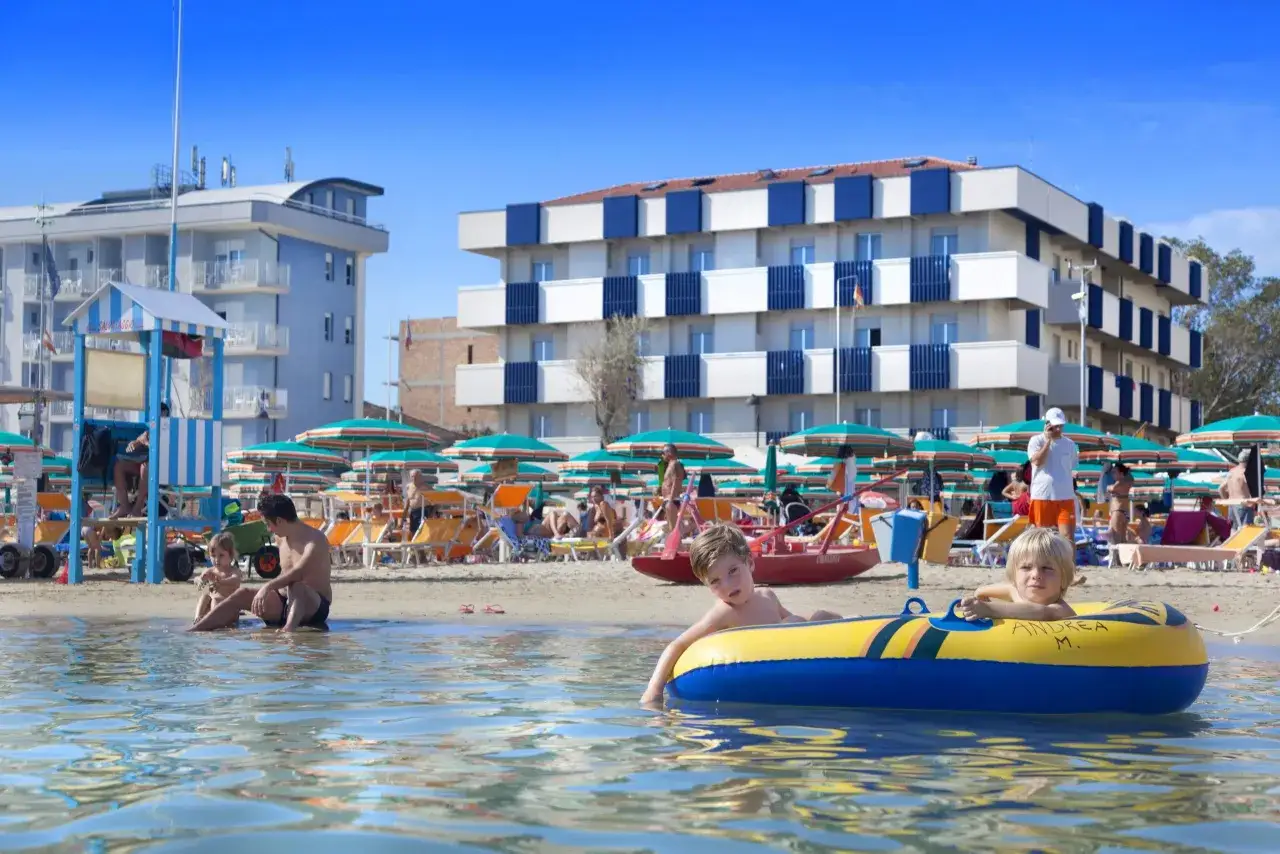 Bambini giocano in acqua davanti al hotel dell'amarissimo, con ombrelloni e lettini sulla spiaggia affollata.