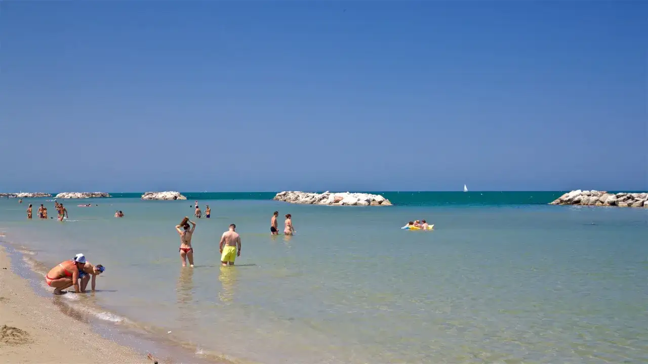 Persone si godono il sole e il mare cristallino sulla spiaggia libera di Torre Pedrera. Un veliero all'orizzonte completa la scena.