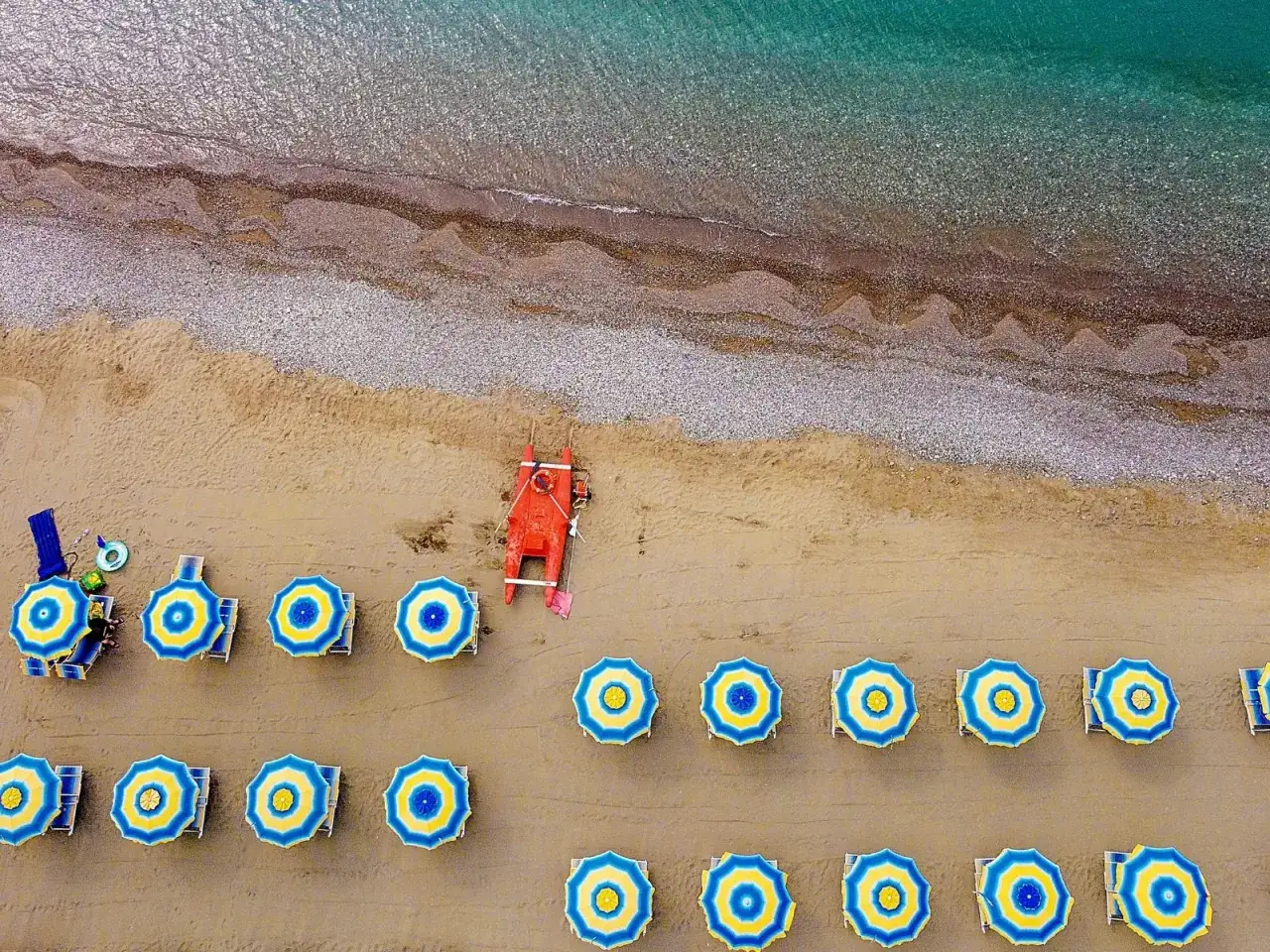 File di ombrelloni blu e gialli e lettini sulla sabbia, con una barca di salvataggio rossa vicino al mare cristallino del parco mare nord Misano.