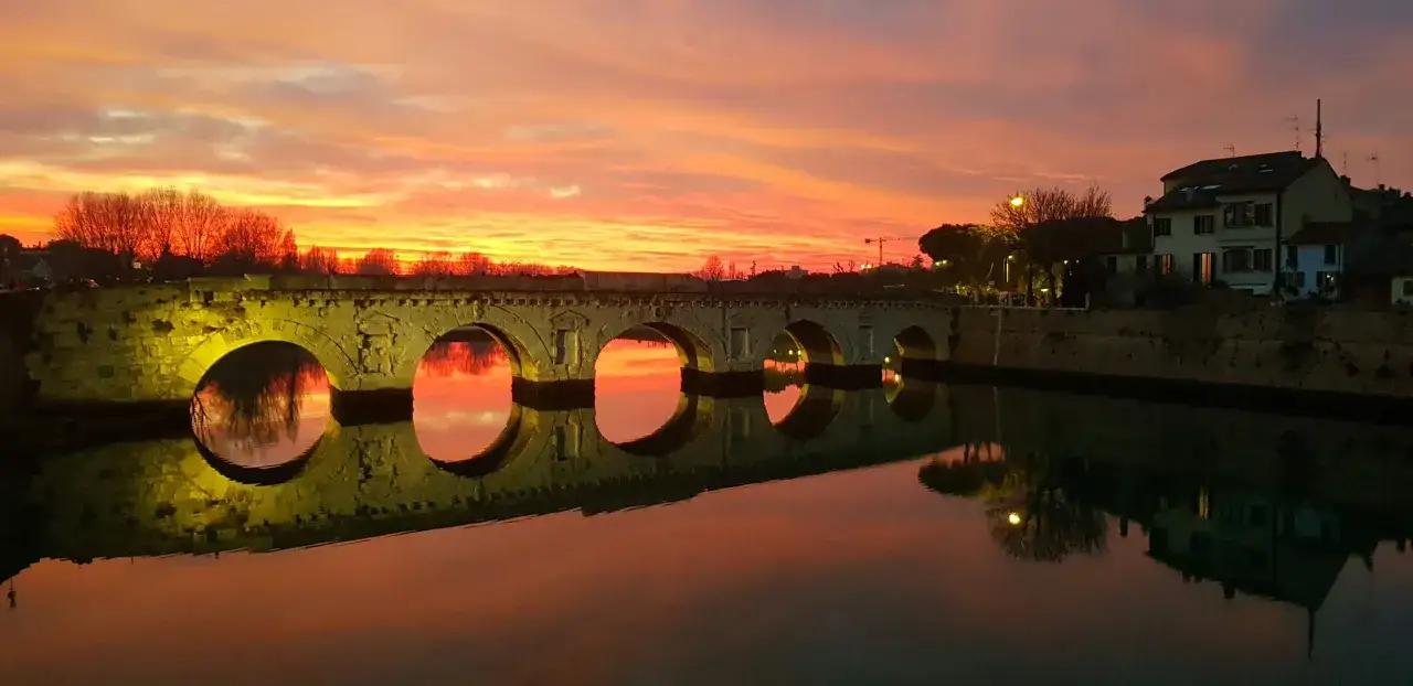 Ponte di Tiberio a Rimini al tramonto, con riflessi sull'acqua e un cielo infuocato. Lo skyline di Rimini è incantevole.