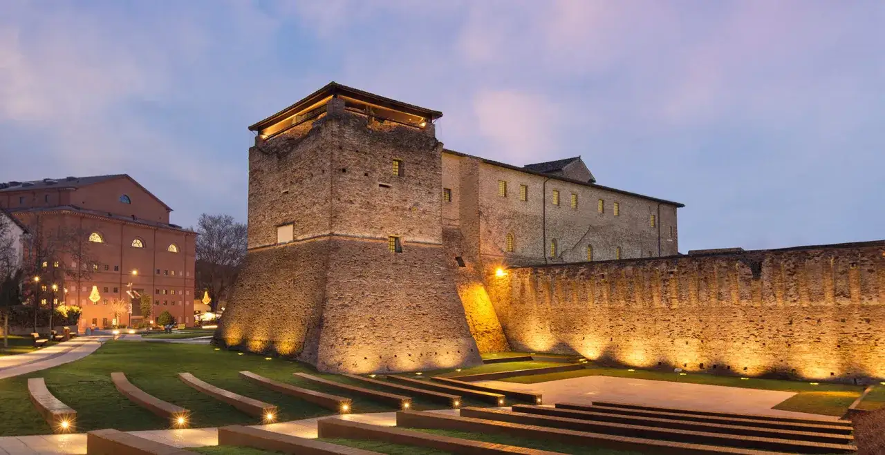 Il Castel Sismondo illuminato di sera, con un parco moderno e un edificio in mattoni rossi sullo sfondo.