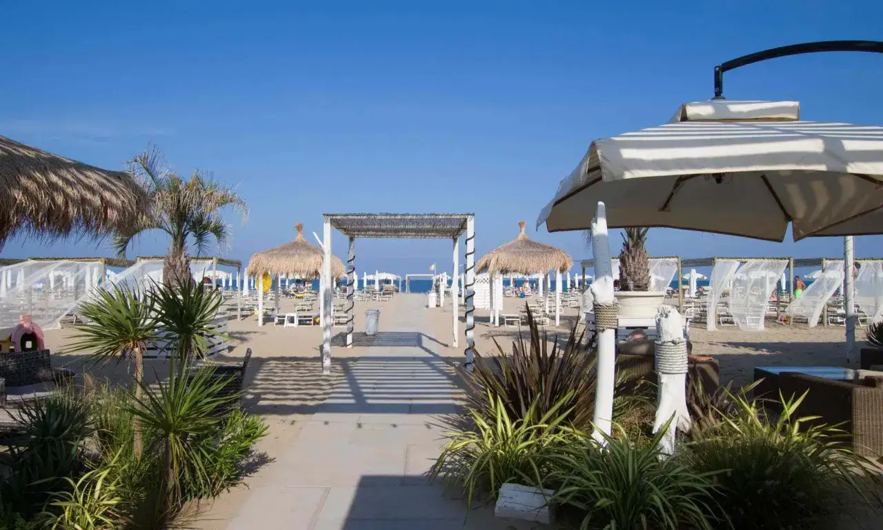 Un percorso conduce al mare a Ravenna, tra ombrelloni di paglia, lettini e tende bianche su una spiaggia sabbiosa.