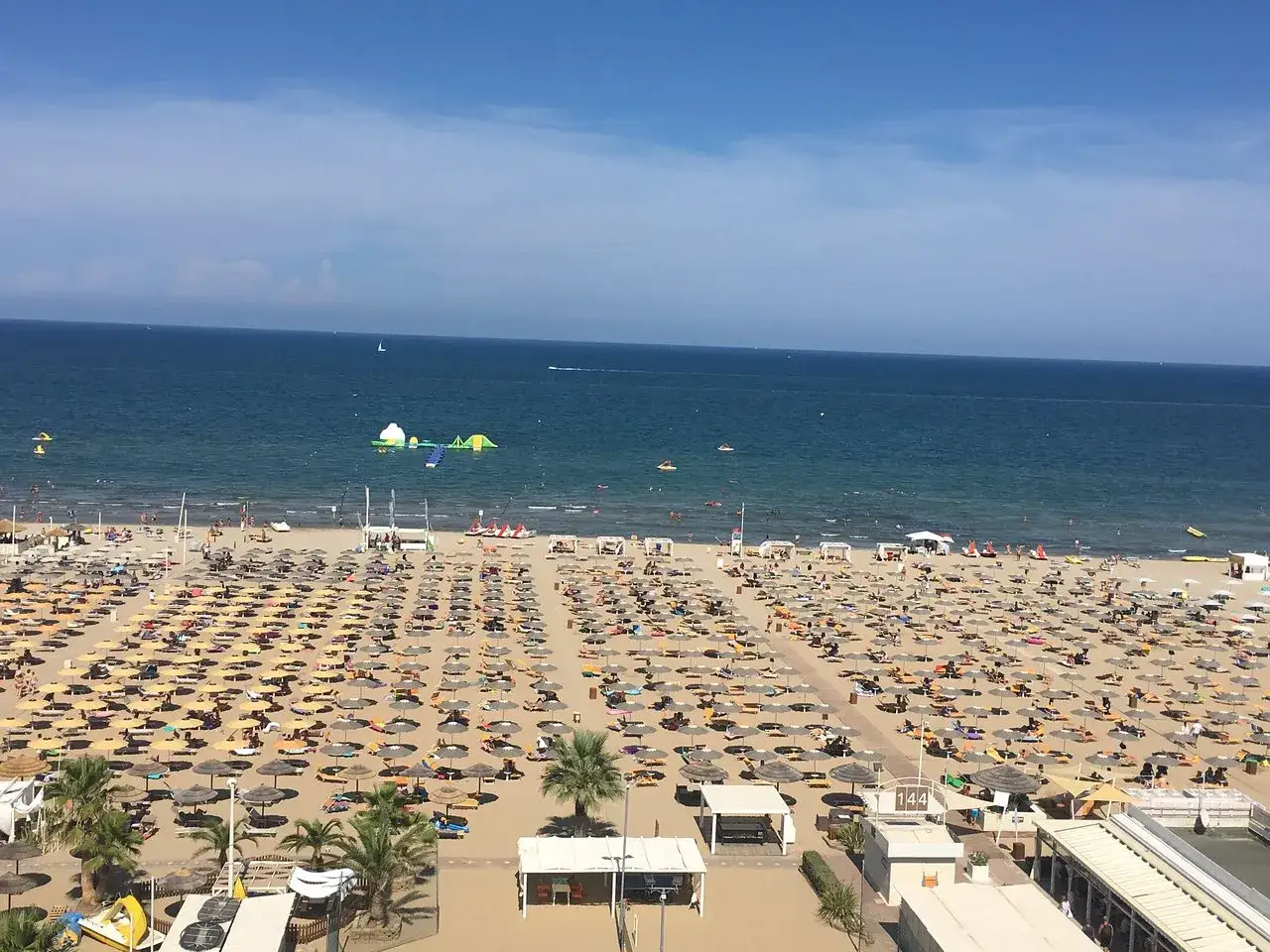 Una vasta spiaggia di Miramare gremita di ombrelloni e lettini, con bagnanti che si godono il mare azzurro e un parco acquatico gonfiabile.