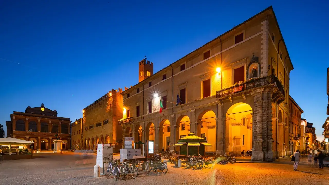 Piazza del Popolo, nel centro di Rimini, illuminata di sera. Edifici storici con portici, biciclette parcheggiate e persone che passeggiano.