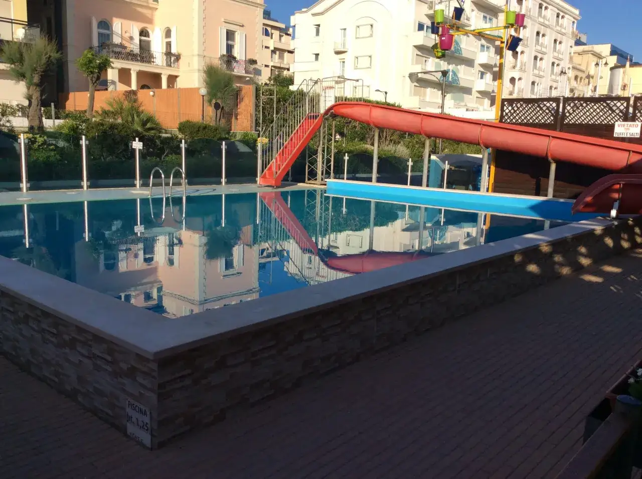Piscina con scivolo rosso e giochi d'acqua ai Bagni Carmen Cattolica. Riflessi degli edifici sull'acqua cristallina.