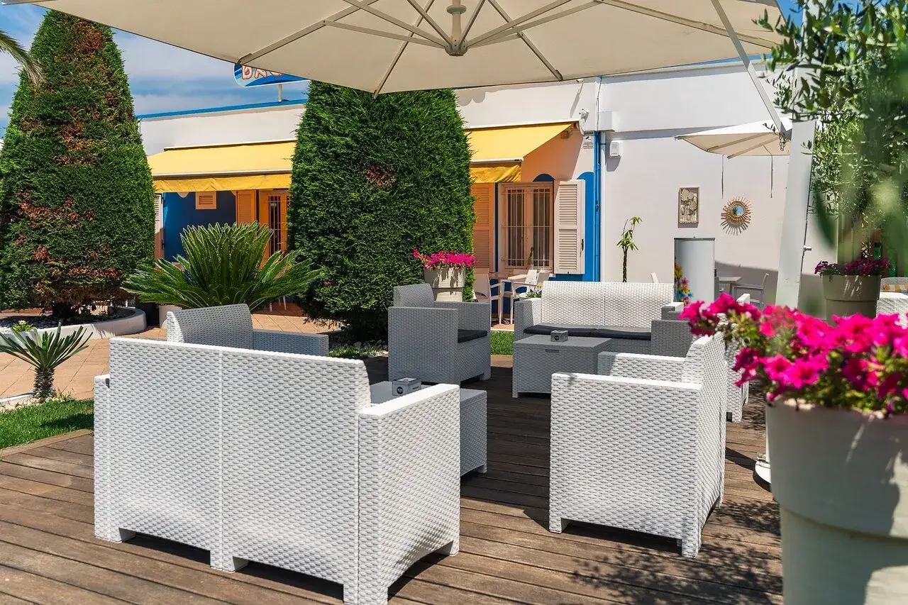 Arredamento da esterno in un bagno giardino a Cesenatico, con divani e poltrone bianche su un deck di legno, circondati da piante verdi e fiori fucsia.