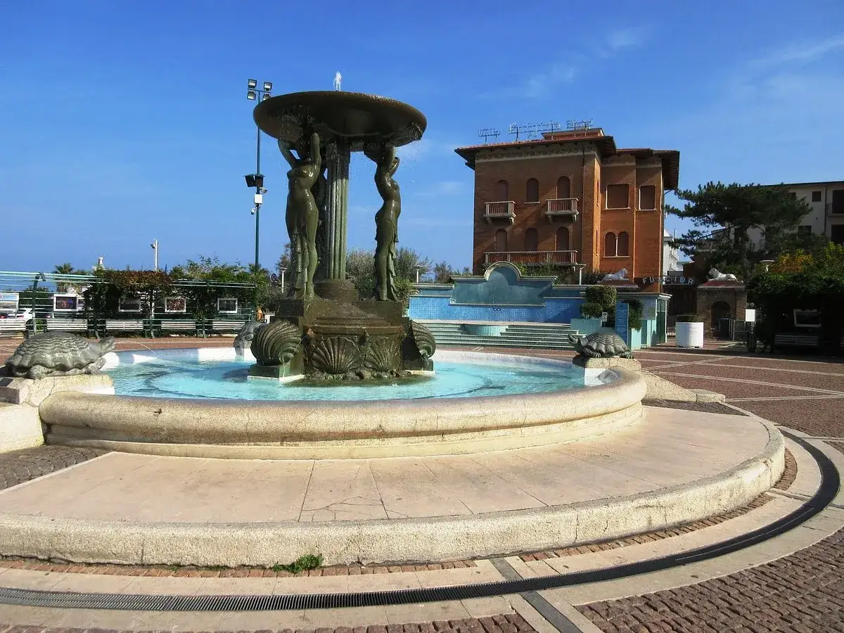 Fontana con statue di donne e tartarughe in piazza Primo Maggio a Cattolica, con un edificio in mattoni sullo sfondo.