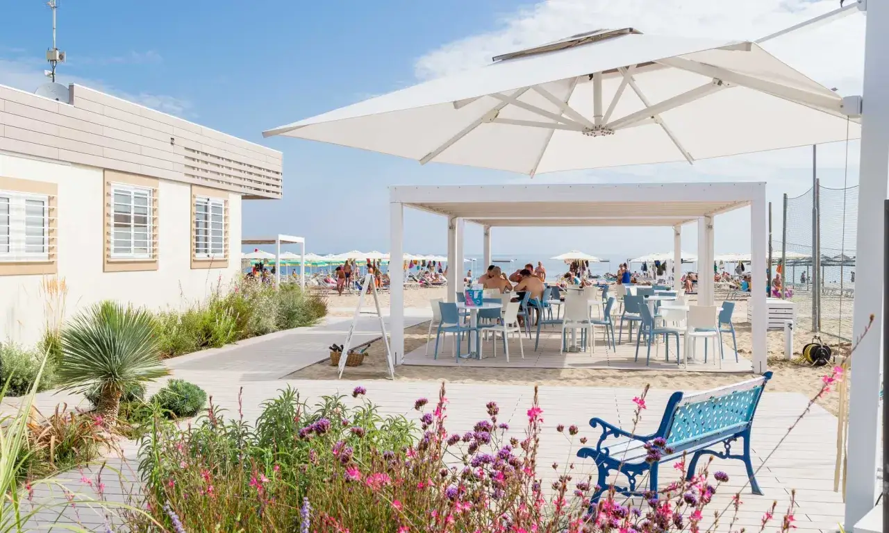 Un angolo di relax a Riccione mare, con tavoli all'aperto sotto un pergolato bianco, fiori colorati e la spiaggia affollata sullo sfondo.