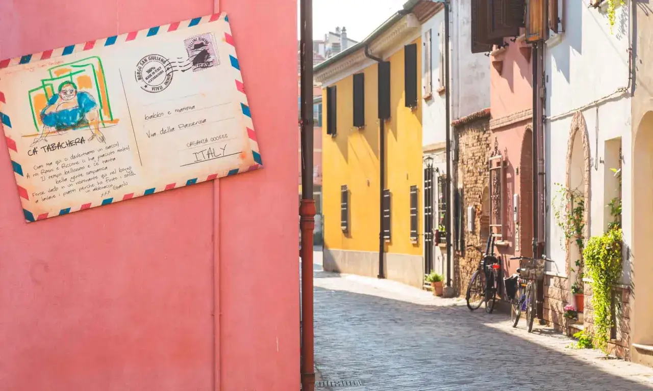 Cartolina da San Giuliano a Mare: un vicolo colorato con bici e case.