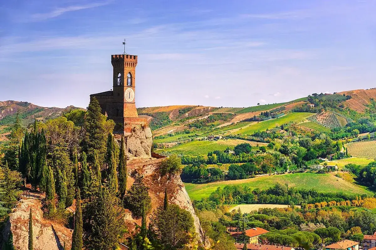Torre medievale su sperone roccioso, circondata da cipressi, domina un paesaggio collinare verdeggiante con vigneti e uliveti, tipico dei borghi dell'Emilia Romagna.