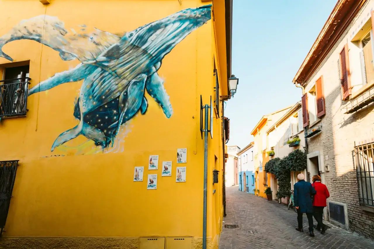 Un murale di balene blu adorna un edificio giallo a San Giuliano Rimini. Una coppia passeggia lungo la strada acciottolata.