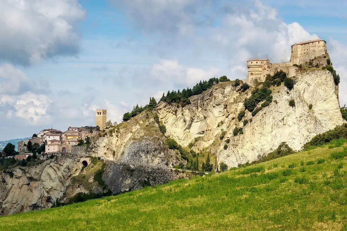 Borgo medievale arroccato su sperone roccioso, con castello e torre, sotto un cielo nuvoloso. Un paesaggio che ricorda la storia, lontano dal mare di Misano Marittima.