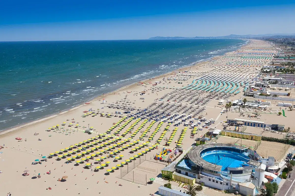 Vasta distesa di spiagge romagna, con file ordinate di ombrelloni gialli e azzurri, bagnanti e una piscina circolare.