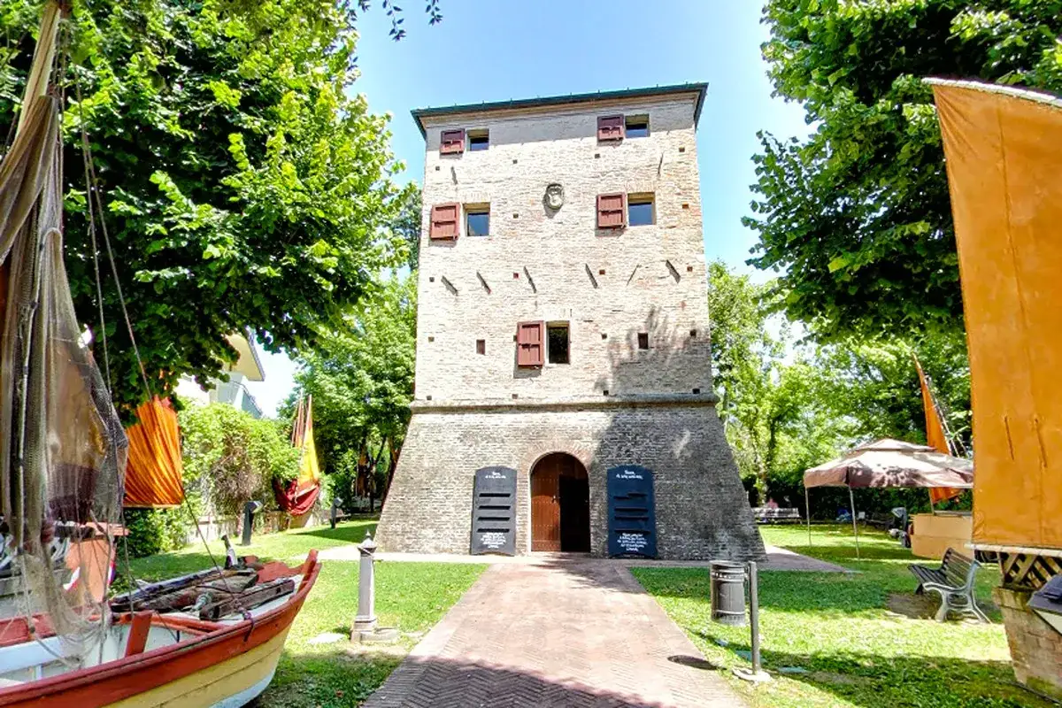Torre antica a Bellaria Igea, con barca e bandiere colorate. Un luogo storico immerso nel verde.