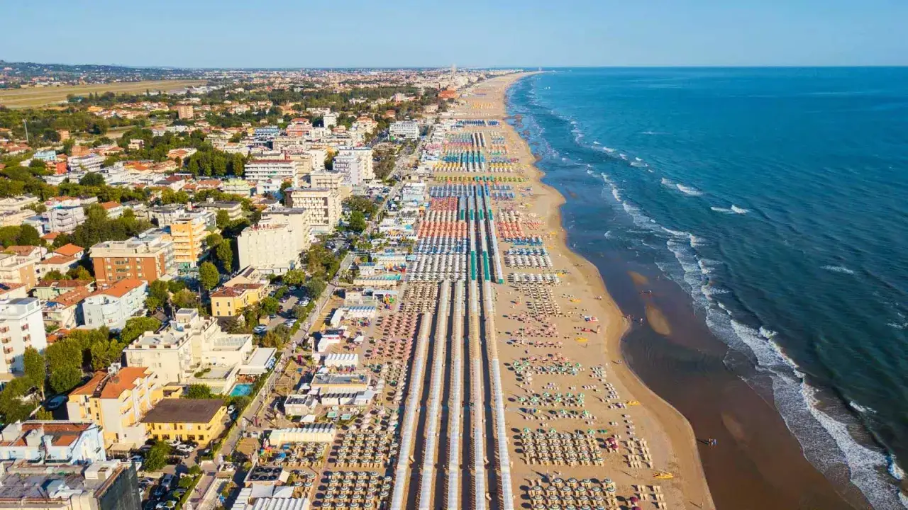 Vista aerea di una lunga spiaggia con file ordinate di ombrelloni e lettini, che si estende fino all'orizzonte. La città di Riccione o Rimini si affaccia sul mare Adriatico.