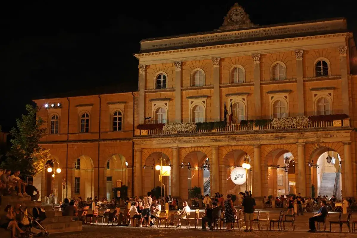 Serata estiva a Rimini Santarcangelo, con gente seduta ai tavolini sotto gli archi illuminati del palazzo storico.