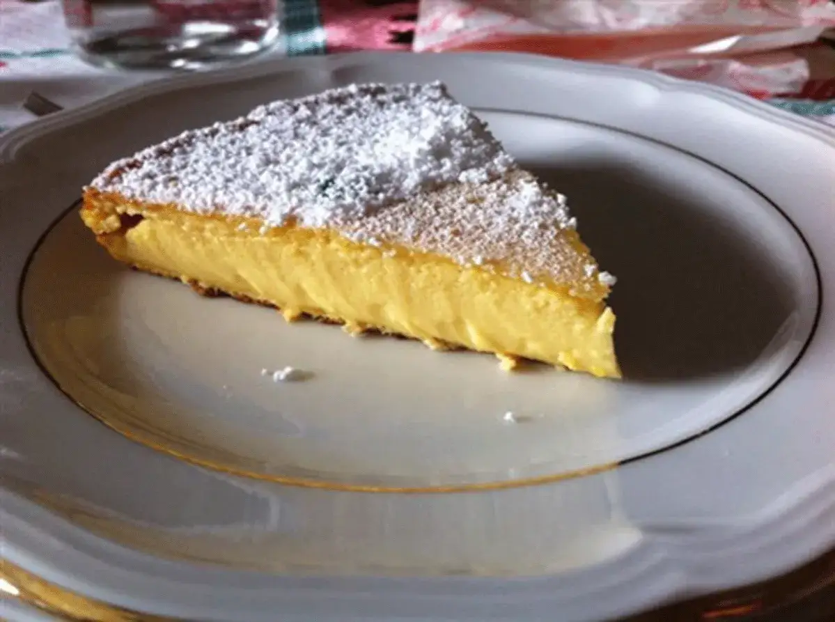 Una fetta di dolce tipico romagnolo, una torta soffice e gialla, cosparsa di zucchero a velo, su un piatto bianco con bordo dorato.