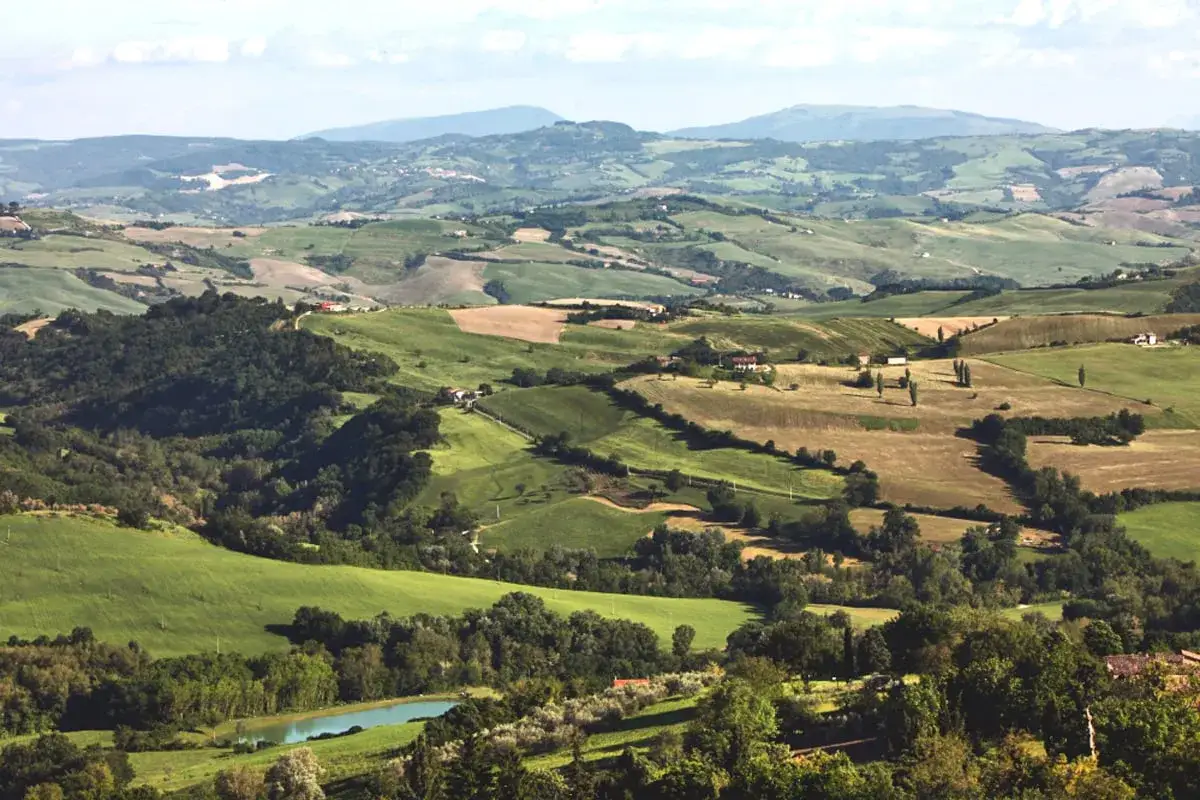 Dolci colline verdi e campi coltivati nella Valconca, Rimini, con un piccolo lago azzurro in primo piano.
