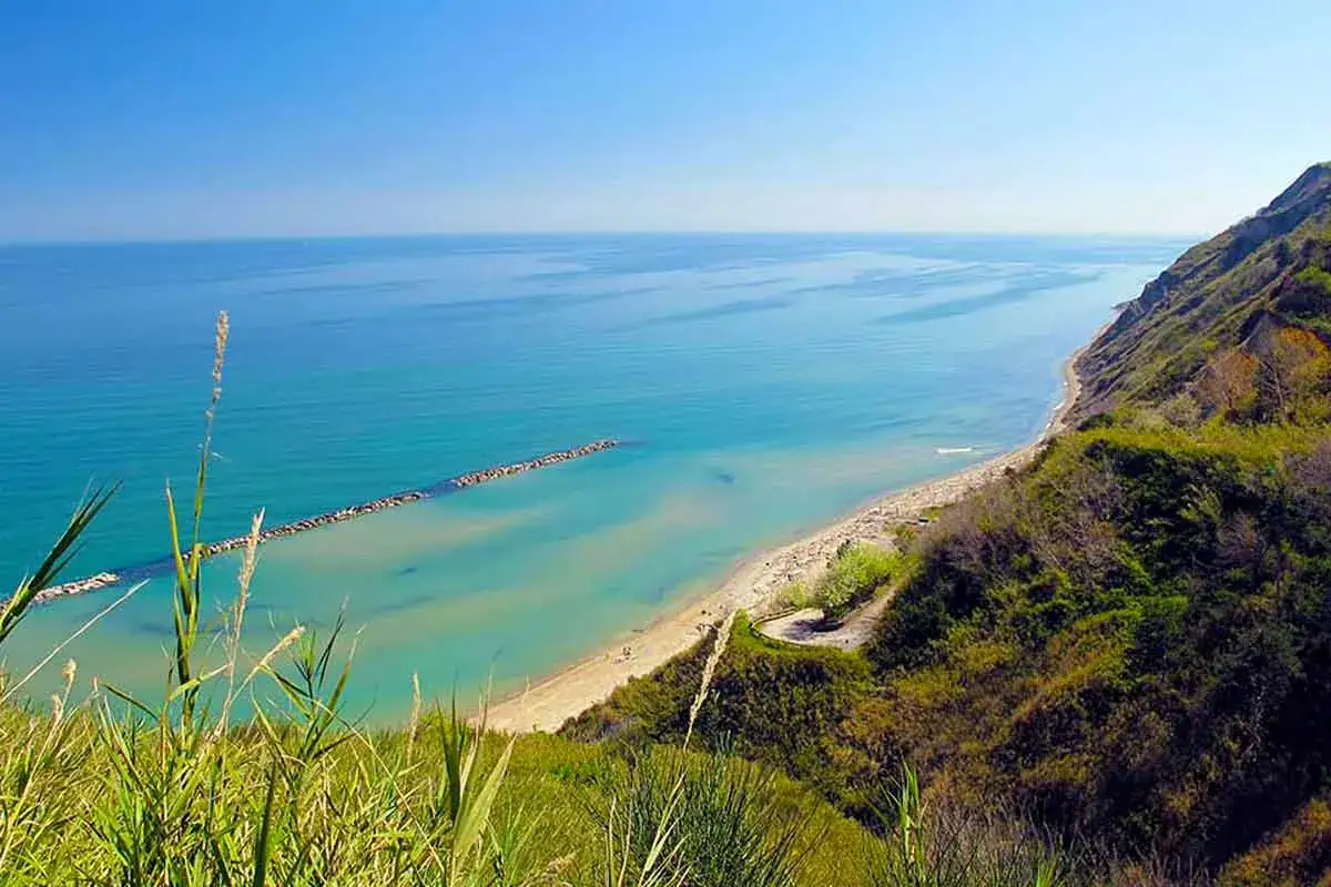 Vista panoramica della spiaggia libera di Gabicce Mare, con mare azzurro, scogliera verdeggiante e vegetazione rigogliosa.