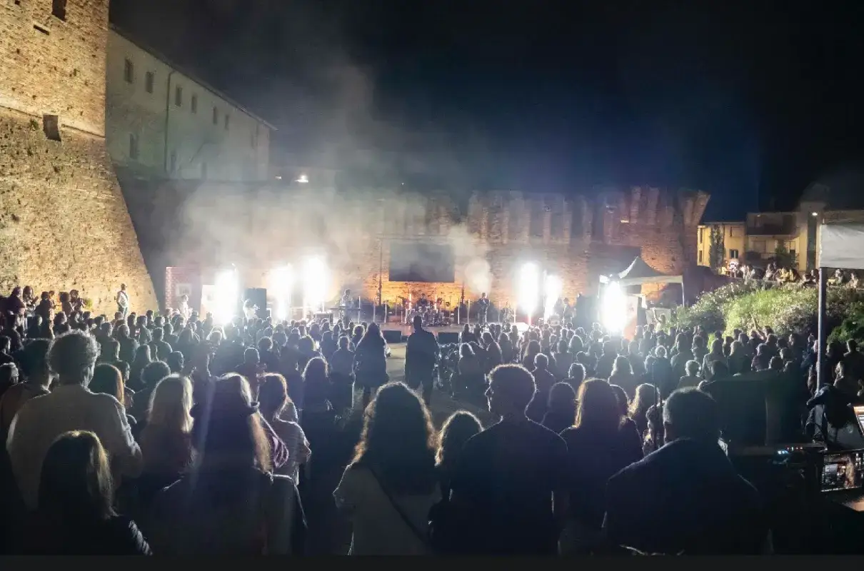 Un grande pubblico assiste a un emozionante concerto rimini, illuminato da potenti fari e fumo scenico, con una band sul palco davanti a un'antica fortezza.