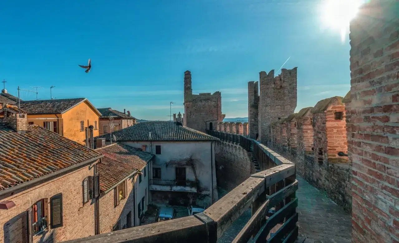 Passeggiata sulle mura di Gradara, cosa vedere: un borgo medievale con torri imponenti, tetti in cotto e un cielo azzurro con un uccello in volo.