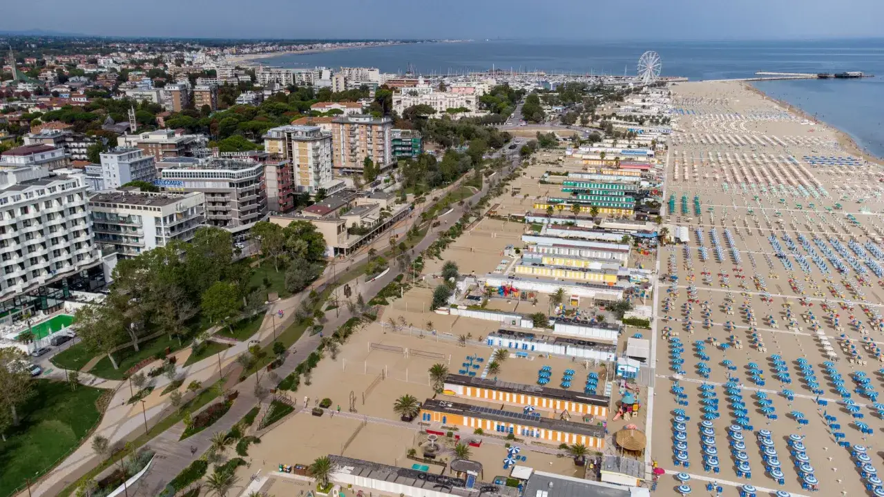 Vista aerea di una vasta spiaggia libera a Rimini, con file ordinate di ombrelloni e cabine colorate che si estendono verso il mare.