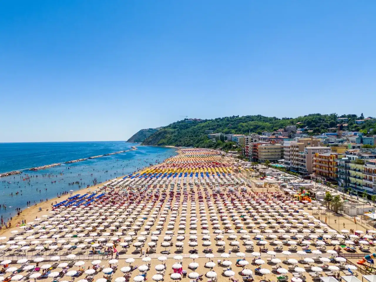 Spiaggia affollata con file infinite di ombrelloni colorati e bianchi. Mare azzurro e collina verde sullo sfondo. Meglio Riccione o Gabicce Mare?