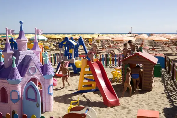 Bambini giocano in un'area giochi colorata sulla sabbia, con castello e scivoli, mentre ombrelloni gialli e lettini creano un'atmosfera da **sunday beach Rimini**.
