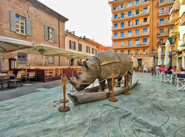 Statua di rinoceronte in una barca, in una piazza di Rimini, dove è nato Fellini. Tavolini all'aperto e palazzi colorati sullo sfondo.