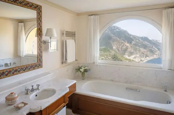 Un bagno di lusso con vasca, lavandino e specchio decorato, che offre una vista mozzafiato sulle montagne e sul mare. Tra i bagni più belli ed eleganti.