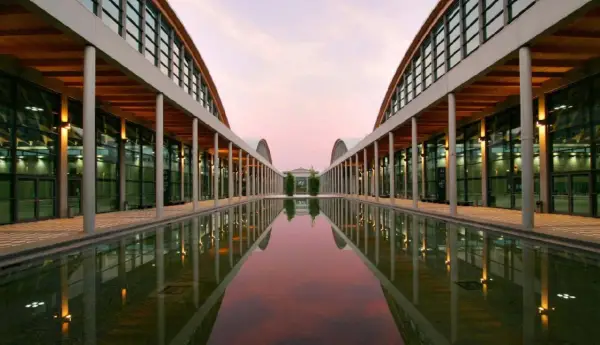 Ingresso moderno della fiera a Rimini, con un lungo specchio d'acqua che riflette le arcate e il cielo rosa del tramonto.