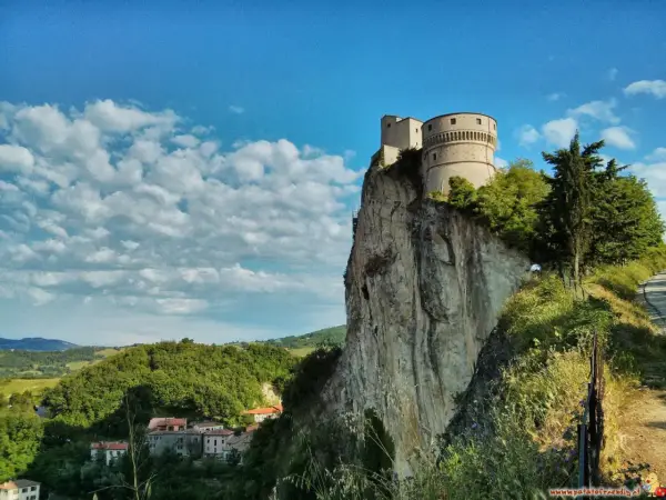 La fortezza di San Leo domina un picco roccioso, con un borgo sottostante e un cielo azzurro punteggiato di nuvole.