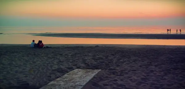 Alba a Rimini: una coppia seduta sulla sabbia guarda il mare calmo e il cielo colorato.