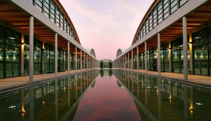 Ingresso moderno della fiera a Rimini, con un lungo specchio d'acqua che riflette le arcate e il cielo rosa del tramonto.